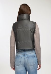 Gilet imbottito grigio scuro tagliato, con colletto alto, indossato sopra un maglione chiaro, abbinato a jeans azzurri chiari con tasche posteriori.