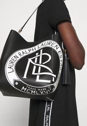 Person in schwarzem Outfit hält große schwarze Tragetasche mit weißem Ralph-Lauren-Logo und Fransen-Detail am Träger.