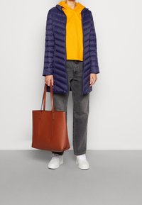 Cappotto imbottito blu navy, felpa gialla con cappuccio, jeans grigi, sneakers bianche e una borsa a mano in pelle marrone con doppi manici. Tessuto texturizzato, design elegante.