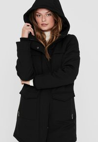 Veste à capuche noire avec fermeture éclair à l'avant. Comprend deux poches avant, un tissu texturé et un ourlet légèrement courbé.