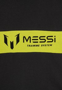 Melnā auduma ar izteiktu dzeltenu horizontālu svītru, uz kuras ir melns teksts: "MESSi TRENIŅU SISTĒMA" un stilizēts logotips virs teksta.