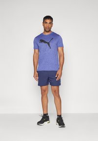 Sinine spordit-särk musta Puma logoga, kombinatsioonis tumepruunide lühikesed püksid ja mustade tossudega. Lihtne disain, lühikesed varrukad, tekstureeritud kangas.