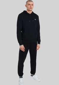 Sudadera negra de algodón con bolsillo frontal, cordones ajustables y un logo. Combinada con joggers negros y zapatillas blancas.