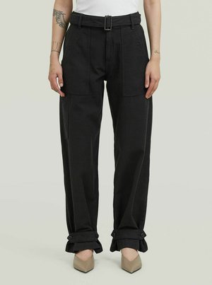 Frau trägt eine schwarze Hose mit hoher Taille und Gürtel, große vorne und an den Seiten Taschen, hochgekrempelte, geknöpfte Ärmelabschlüsse, beige spitze Schuhe und ein weißes Oberteil.