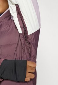 Vijolična puhasta jakna z belim zgornjim delom. Ima stranski zadrgo in elastične črne manšete. Teksturiran, lahek material.