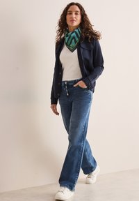 Marineblå knapjakke over en hvid top, parret med lyseblå bukser med brede ben og hvide sneakers. Et grønt mønstret tørklæde er om halsen.