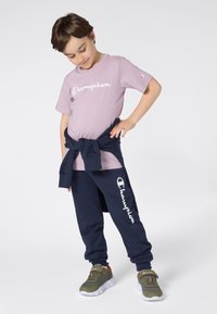 Camiseta lila con el logo de "Champion", combinada con pantalones deportivos azul marino con estampado de logo. Zapatillas deportivas grises con acentos verdes completan el look.