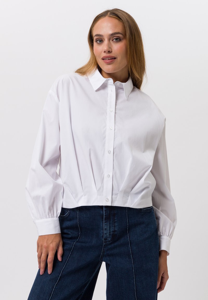 Witte cropped blouse met knoopsluiting aan de voorkant, een spreidkraag en ballonmouwen. Gladde stof, minimalistisch ontwerp. Gecombineerd met donkerblauwe spijkerbroek.