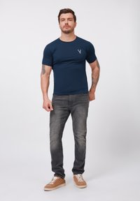 Camiseta de manga corta azul marino con un pequeño emblema de ciervo, combinada con jeans grises y zapatillas de color beige claro. Diseño simple con una forma ajustada.