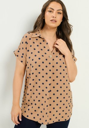 Kvinde med langt mørkt hår har en beige kortærmet bluse med sorte polkaprikker og en enkelt knap nær kraven.