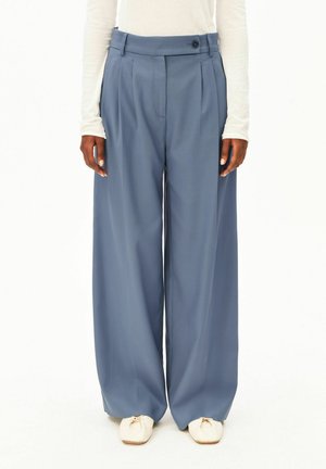 Femme portant un pantalon bleu taille haute à jambes larges, un haut blanc à manches longues et des chaussures blanches à bout carré, debout devant un fond blanc.