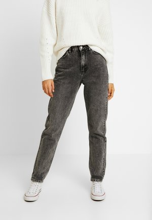 Personne portant un jean noir délavé à coupe ample, un pull en maille blanc et des baskets blanches, debout avec les mains détendues le long du corps.