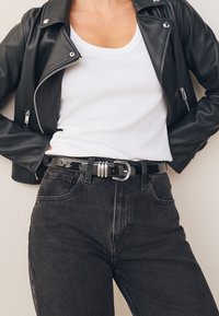 Veste de moto en cuir noir avec des accessoires argentés, associée à un débardeur ajusté blanc et un jean noir taille haute avec une ceinture noire brillante.