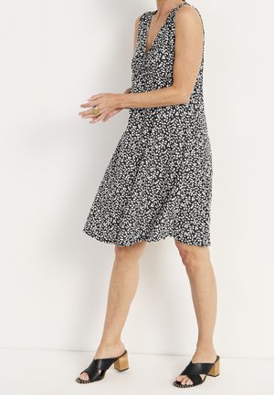 Kobieta w czarnej sukience bez rękawów z białymi wzorami liści oraz czarnych sandałach na drewnianym klocku, stojąca przy białej ścianie.