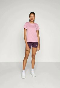 Lys rosa treningstopp med korte ermer og mørk lilla shorts, begge med små logoer. Modellen har på seg hvite sneakers og sokker.