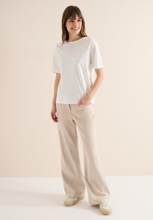 Lächelnde Frau steht und trägt ein weißes Kurzarmhemd, beige weit geschnittene Hosen und weiße Turnschuhe vor einem einfarbigen beigen Hintergrund.