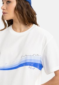 Vit bomull t-shirt med en blå grafisk design som har tre horisontella ränder och en strandillustration nära halsringningen.
