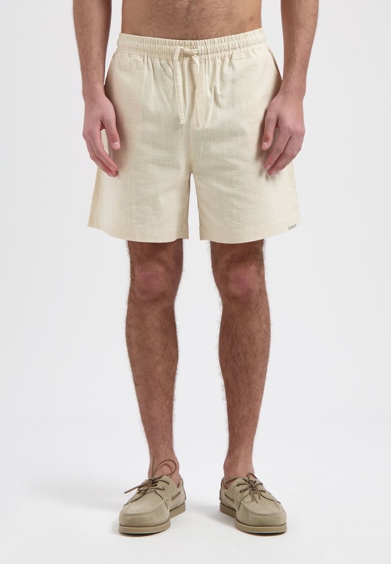 Beige linnen shorts met een elastische tailleband en trekkoord, voorzien van een gestructureerd oppervlak en zijzakken, gecombineerd met lichte schoenen.