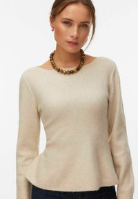 Femme portant un pull à manches longues beige clair, un pantalon foncé, un collier en perles marron et des petites boucles d'oreilles anneaux sur fond blanc.