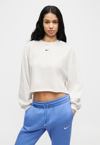 Γκρι cropped sweatshirt με μακριά μανίκια και μαύρο λογότυπο Nike· συνδυάζονται με μπλε φόρμα με ελαστική μέση και λευκό λογότυπο.