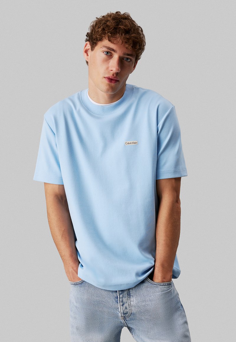 T-shirt in cotone azzurro chiaro con maniche corte e scollatura a girocollo. Presenta un piccolo logo beige di Calvin Klein sul petto. Indossato con jeans di denim chiaro.
