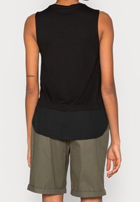 Černý top bez rukávů s vrstveným designem na zadní straně, spárovaný s khaki šortkami. Tkanina vypadá hladce, má volný střih a jednoduché okraje.