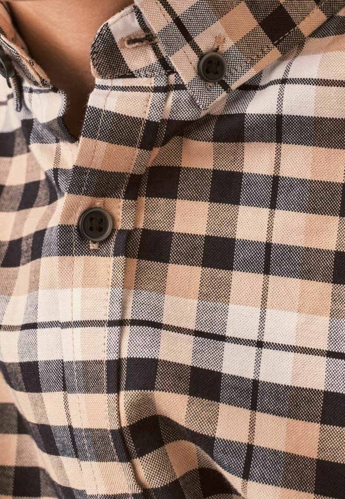 Gros plan sur une chemise à carreaux beige, noire et blanche avec deux boutons noirs visibles, portée par une personne.
