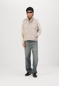 Lichtbeige puffer vest met een hoge kraag en horizontale stiksels, gecombineerd met een grijze langekledingtop en blauwe jeans, gedragen met zwarte sneakers.