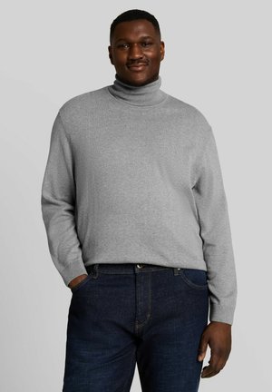 Man with dark skin smiling, wearing a light gray turtleneck sweater and dark blue jeans, standing against a plain background.