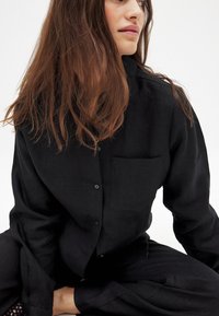 Black button-up shirt made of textured fabric, with a single chest pocket and loose fit, paired with matching pants, displayed against a light background.