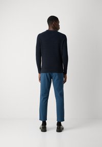 Marineblauer gerippter Strickpullover kombiniert mit blauen Baumwollhosen, von hinten gezeigt. Verfügt über lange Ärmeln und eine taillierte Passform, ergänzt durch schwarze Schuhe.