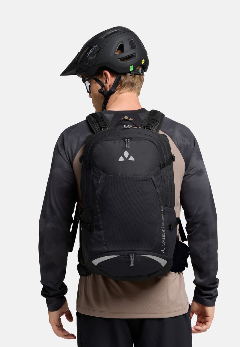 Zaino nero con tessuto testurizzato e molteplici scomparti, indossato con un casco nero. Presenta dettagli riflettenti e spallacci regolabili.