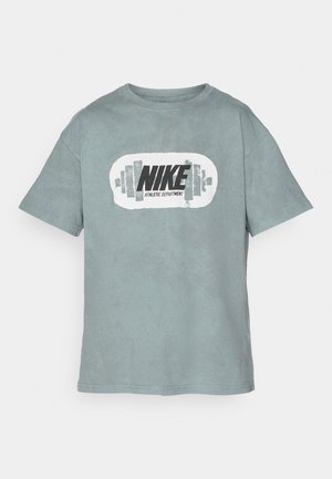 Hele-sinine puuvillane T-särk, millel on ümmargune mustvalge graafika, kus on kirjas "NIKE" ja "ATLETI OSAKOND" ning kangist sümbolid.