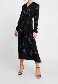 Robe longue noire à manches longues avec un motif floral bleu et orange, col, boutonnière à l'avant et une ceinture à nouer à la taille, accompagnée de talons noirs.