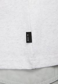 Witte, gestructureerde stof t-shirt met een zwart etiket met de tekst "MAX 90." De onderzoom is iets langer en heeft een lichtgrijze voering aan de onderkant.
