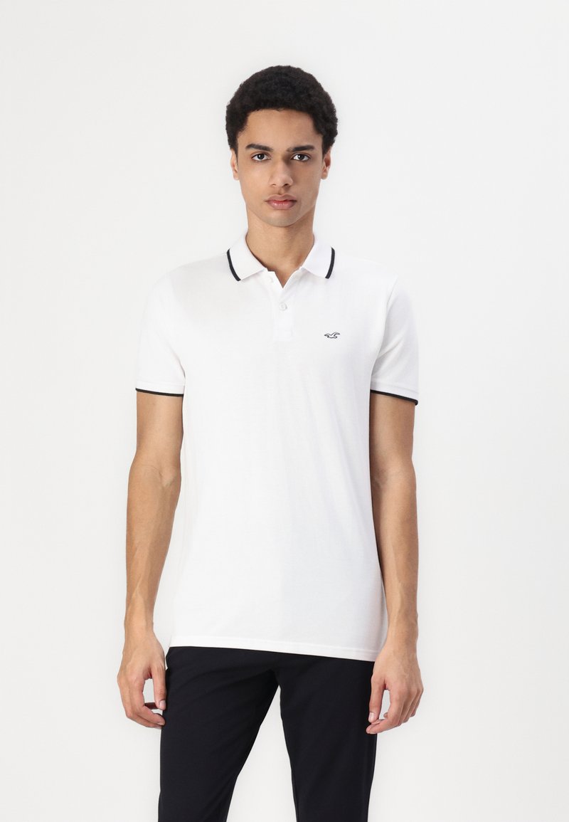 Polo shirt blanc avec des accents noirs sur le col et les manches, fabriqué en tissu lisse. Présente un petit logo sur la poitrine gauche.