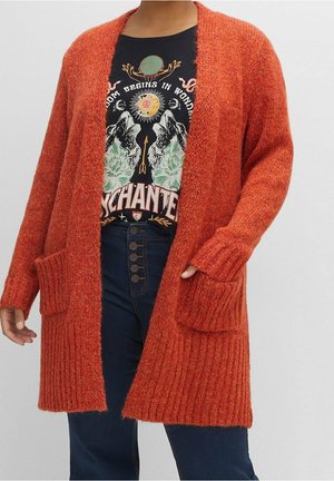 Personne portant un long cardigan en tricot orange avec des poches, un t-shirt noir à motif graphique et un jean bleu taille haute avec des boutons.