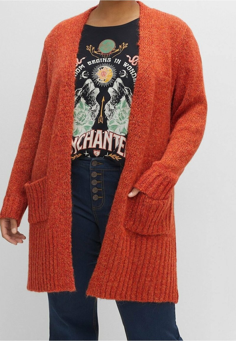 Personne portant un long cardigan en tricot orange avec des poches, un t-shirt noir à motif graphique et un jean bleu taille haute avec des boutons.