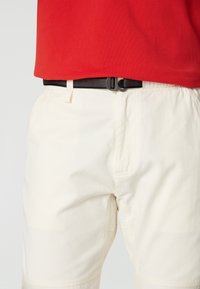 Vita shorts med en texturerad yta, utrustade med ett svart mesh-bälte med en rund spänne, tillsammans med en röd skjorta.