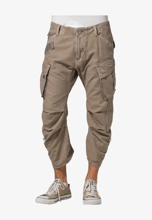 Pantalon cargo court marron clair avec plusieurs poches porté par une personne debout, associé à des baskets basses beige clair.