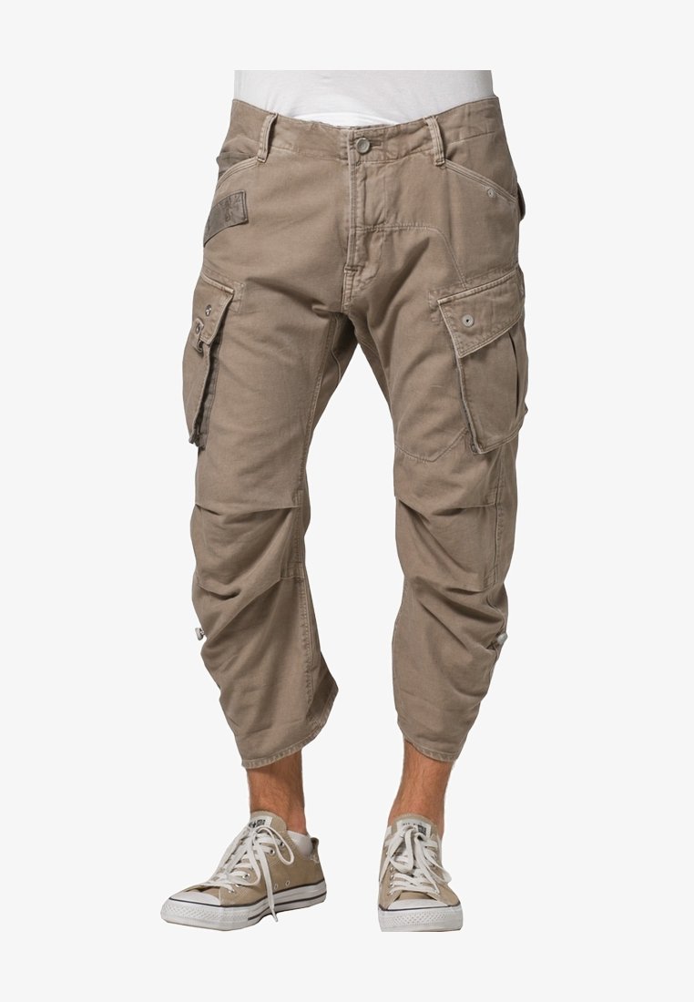 Pantalon cargo court marron clair avec plusieurs poches porté par une personne debout, associé à des baskets basses beige clair.