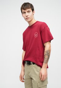 Νεαρός άνδρας σε κόκκινο γραφικό μπλουζάκι και μπεζ καλοκαιρινό παντελόνι στέκεται με το τατουάζ στους καρπούς του ορατό, μπροστά από έναν απλό λευκό τοίχο.