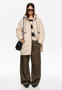 Abrigo beige acolchado con diseño de cuadros y capucha, usado sobre un suéter de rayas negras y crema, combinado con pantalones de pierna ancha en verde oliva.