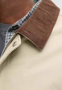 Beige jakke med en brun corduroy-krave, accentueret med et ternet flannel-foring. Glat stof med en knaplukning.