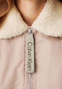 Beige bunda s načechraným krémovým límcem, středovým stříbrným zipem a béžovou táhlicí na zip označenou "Calvin Klein."