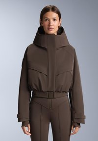 Ruskea pehmustettu takki korkealla kauluksella, jossa on suuret etutaskut ja vyöllinen vyötärö. Suora rakenne ja sileä pinta.