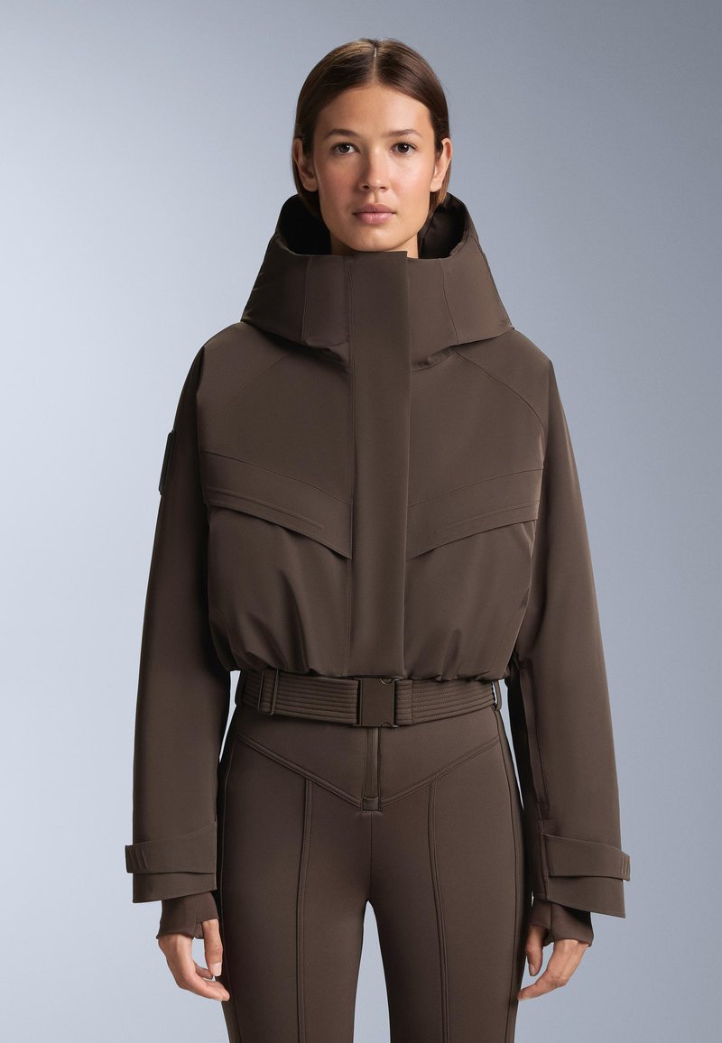 Hnedá prešívaná bunda s vysokým golierom, obsahujúca veľké predné vrecká a opasok v páse. Hladká textúra a štruktúrovaný dizajn.