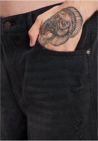 Jeans de denim negro con un primer plano de un detalle de bolsillo, mostrando tela desgastada con pequeños desgarros y un acento de botón metálico visible.