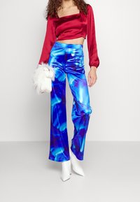 Žena v lesklých modrých kalhotách s širokými nohavicemi, červeném saténovém topu ke krku, bílých kotníkových botách a držící bílo-srstou kabelku.