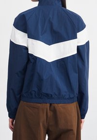 Námorně modrá větrovka s bílými chevronovými pruhy na zádech, s elastickým lemem a manžetami, vyrobená z lehkého, vodoodpudivého materiálu.